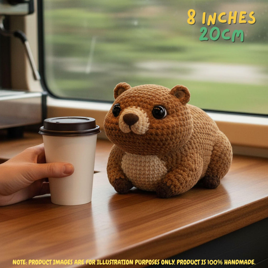 A crocheted brown bear plush toy sits on a wooden table next to a paper coffee cup held by a hand, with a blurred window background and text reading '8 INCHES 20CM'.