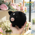 A black crocheted pig toy being held in a person's hand, with pink flowers and a window in the blurred background. Yellow text in the top right reads '6 INCHES 16CM'.