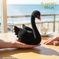 A crocheted black swan toy with a red and white beak is held in two human hands on a wooden table, with a beach background and text indicating 6 inches (16 cm) size.