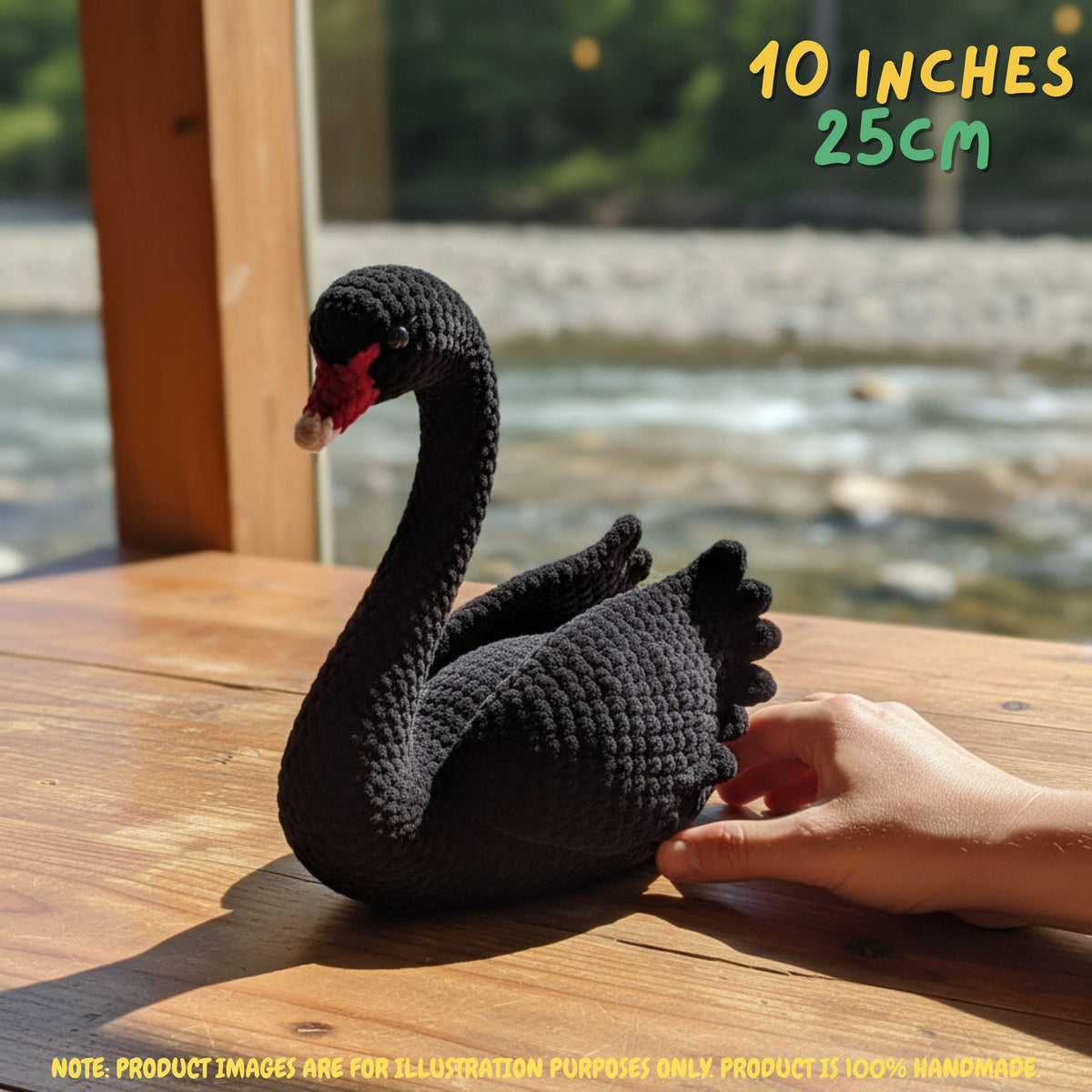 A crocheted black swan figurine with a red beak and white tip, displayed on a wooden table with a hand supporting it. A blurred outdoor river scene is in the background, with text overlays indicating the size '10 INCHES 25CM'.