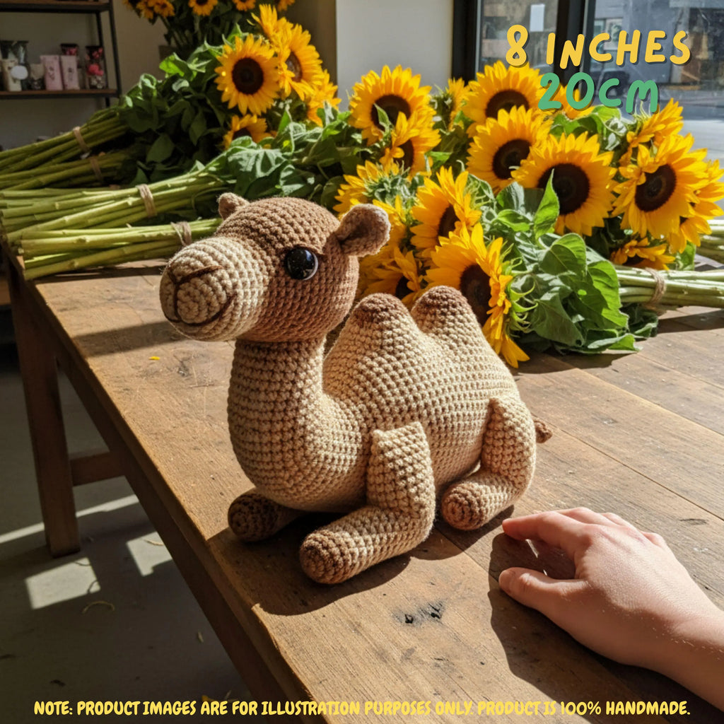 A crocheted camel toy sits on a rustic wooden table, with a person's hand reaching toward it. Bright sunflowers are arranged in the background, and text overlays indicate the size as 8 inches (20cm) with a note that the product images are for illustration purposes only since the product is 100% handmade.