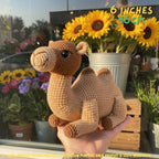 A crocheted camel toy being held in a person's hand in front of a window with sunflowers and flowers outside. Yellow text in the top right says '6 INCHES 16CM'.