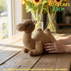 A crocheted camel toy sits on a wooden table with a person's hands touching it for scale. The background shows a blurred sunflower arrangement in a vase near a window.