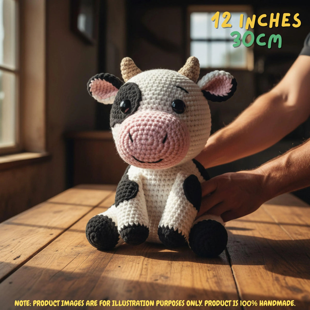 A crocheted cow plush toy sits on a wooden table in warm natural light, with a person's arm reaching toward it. Text in the top right reads '12 INCHES 30CM', and a note at the bottom says the product image is for illustration purposes only and the product is 100% handmade.