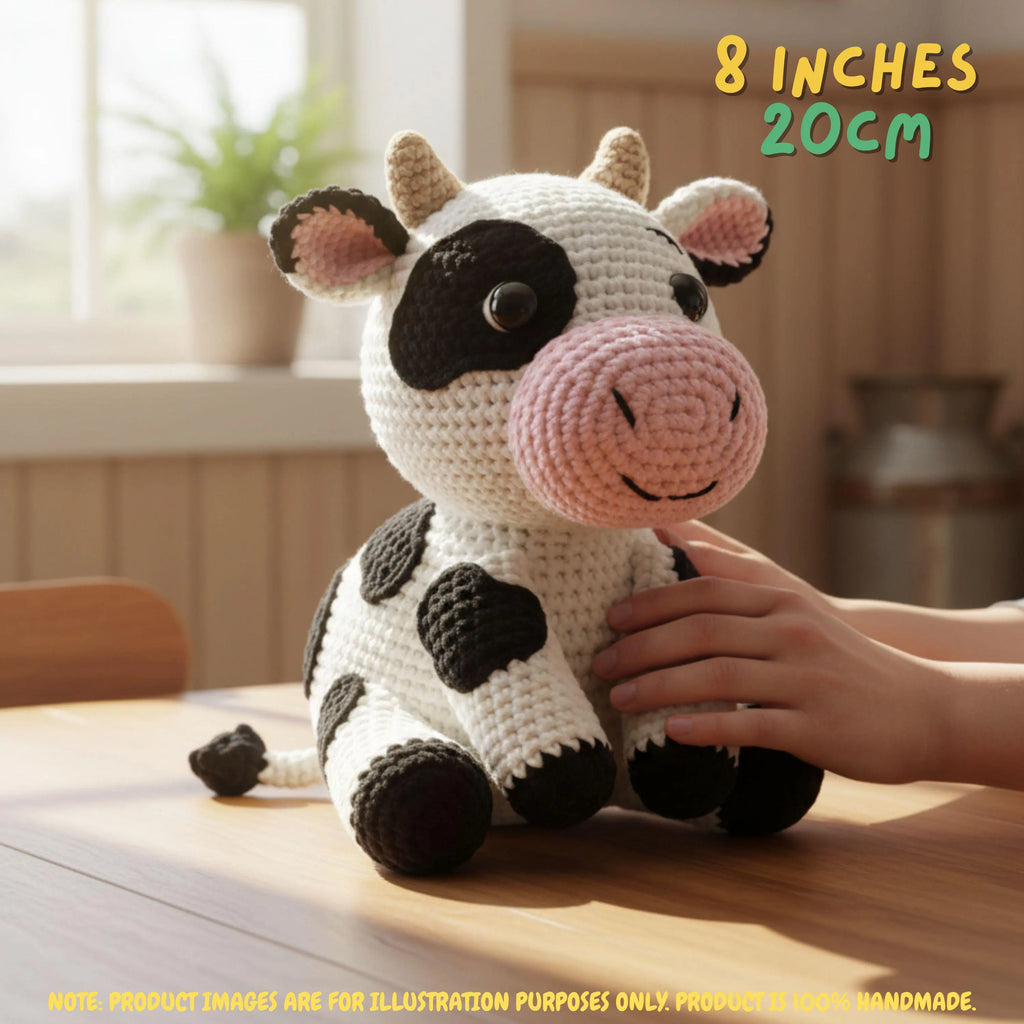 A crocheted cow plush toy sits on a wooden table in a warm indoor setting, with a human hand resting on its side. The toy is white with black spots and a pink snout and inner ears.