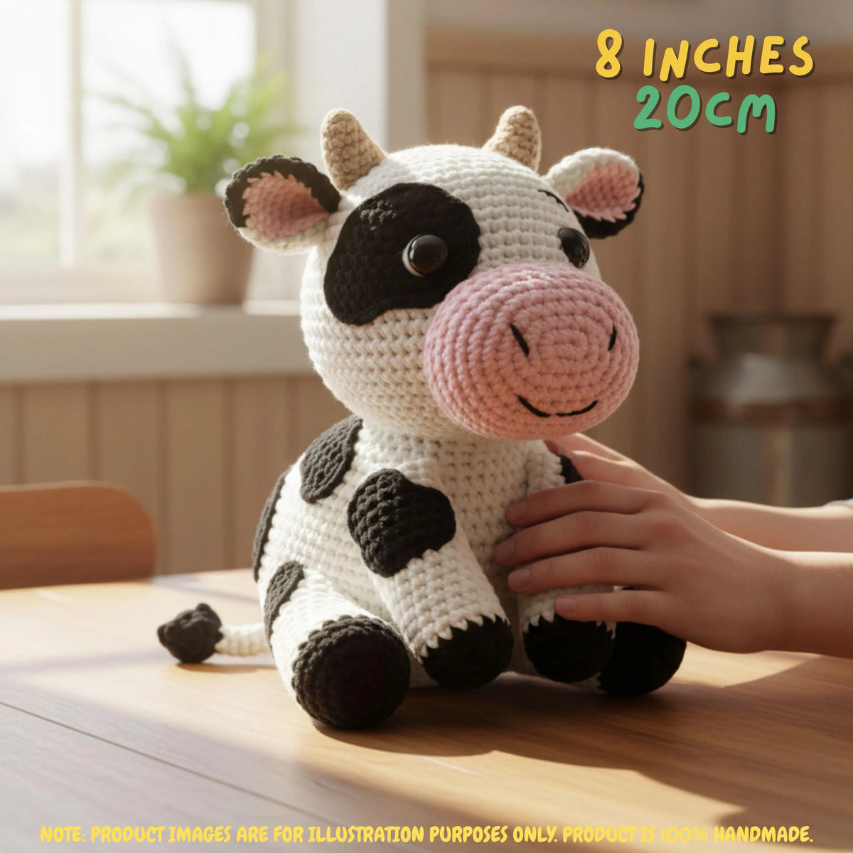 A crocheted cow plush toy sits on a wooden table in a warm indoor setting, with a human hand resting on its side. The toy is white with black spots and a pink snout and inner ears.