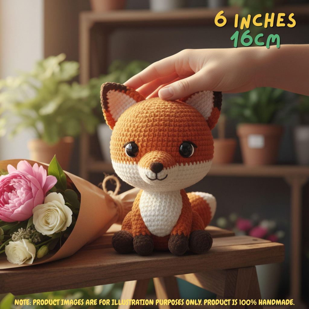 A crocheted plush fox toy sits on a wooden surface in a warm indoor setting, with a human hand gently touching its head. The fox is orange and cream with dark brown paws, feet, and inner ears.