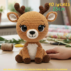 A crocheted stuffed deer toy sits on a tabletop in a real indoor setting, with a human hand touching it and decorative items blurred in the background. Text in the image reads '10 INCHES 25CM' and there is a note at the bottom saying the product image is for illustration purposes only and the product is 100% handmade.
