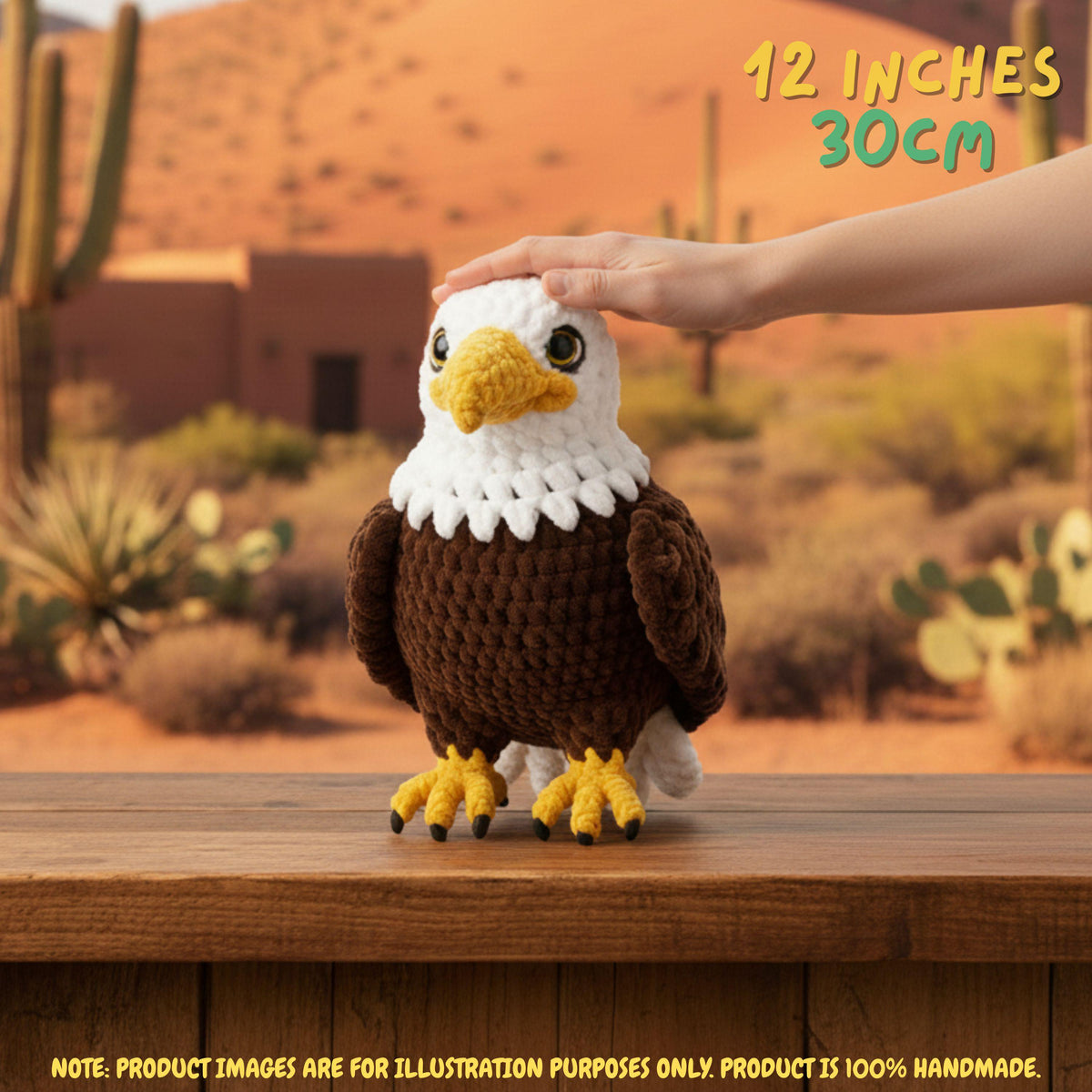 A crocheted eagle toy sits on a wooden table in a desert landscape setting, with a human hand gently resting on its head. The text '12 INCHES 30CM' is visible in the top right corner.