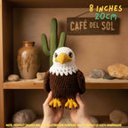 A crocheted eagle toy held up by two hands in front of a rustic wooden shelf display. The text overlay reads '8 INCHES 20cm'.