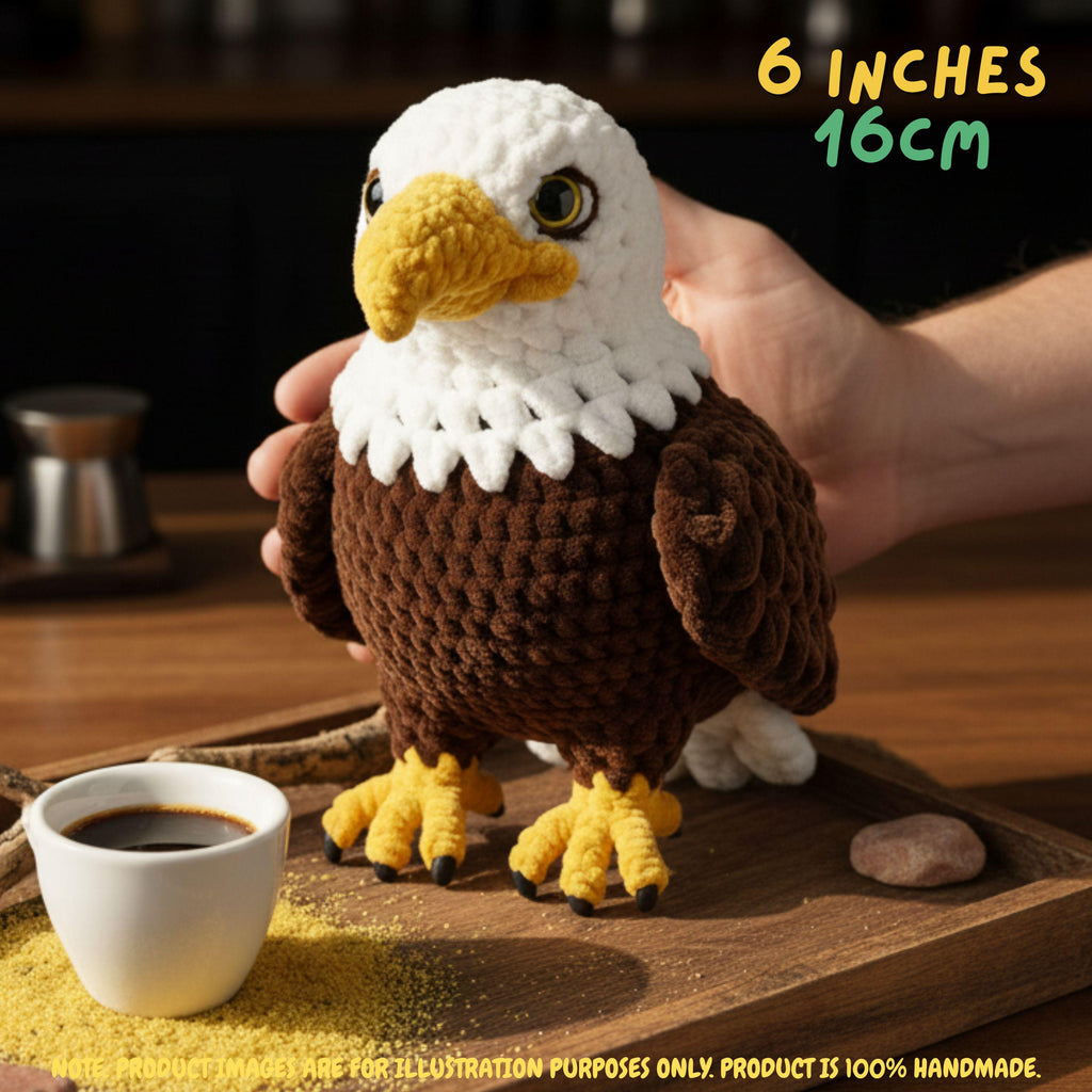 A handmade crocheted eagle plush toy being held by a person's hand on a wooden table, with a small cup of coffee and yellow cornmeal nearby. Text in the top right reads '6 INCHES 16CM'.