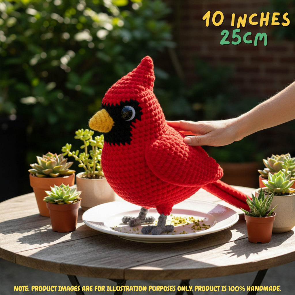 A red crocheted cardinal bird toy standing on a white plate on a wooden table outdoors, with a human hand touching its back. Small potted succulents surround it, and text in the top right says 10 INCHES 25CM.