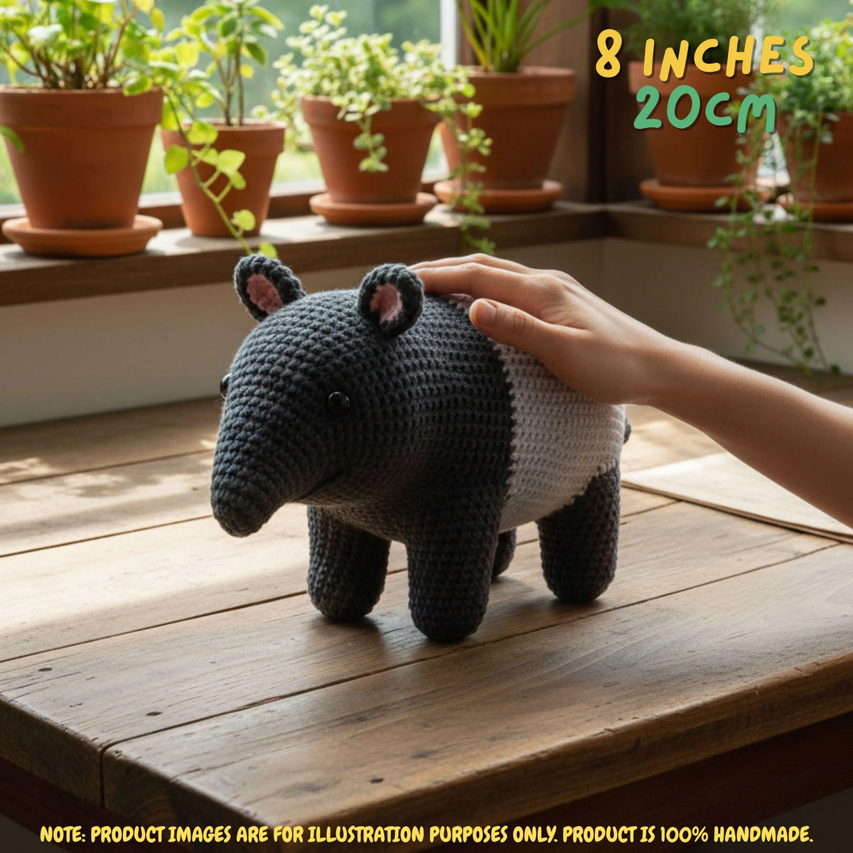 A crocheted armchair shaped like a small animal, shown on a wooden table with a human hand resting on its back. The chair is dark charcoal gray with a white side panel and pink inner ears.
