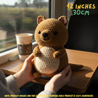 A crocheted plush kangaroo toy being held in two hands on a wooden table near a window. The toy is brown with a cream-colored belly and small pink inner ears.