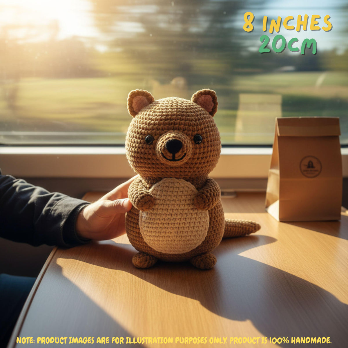 A crocheted plush bear toy sits on a wooden table by a window, with a human hand touching its side. The text '8 INCHES 20CM' appears in the top right, and a small brown paper bag is visible to the right.
