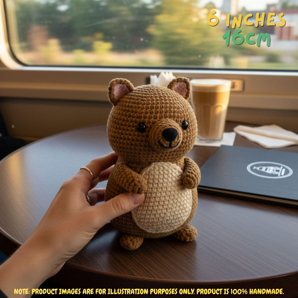 A small crocheted teddy bear plush toy is being held by a person's hand on a wooden table near a window. The bear has a medium brown body, a cream-colored belly, and black embroidered eyes and nose.