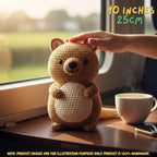 A crocheted kangaroo toy sits on a wooden table by a window, with a human hand resting on its head. A white mug and phone are beside it, and text in the image notes 10 inches / 25 cm.