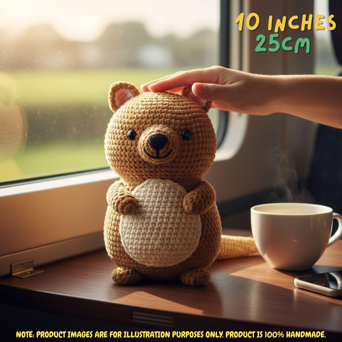 A crocheted kangaroo toy sits on a wooden table by a window, with a human hand resting on its head. A white mug and phone are beside it, and text in the image notes 10 inches / 25 cm.