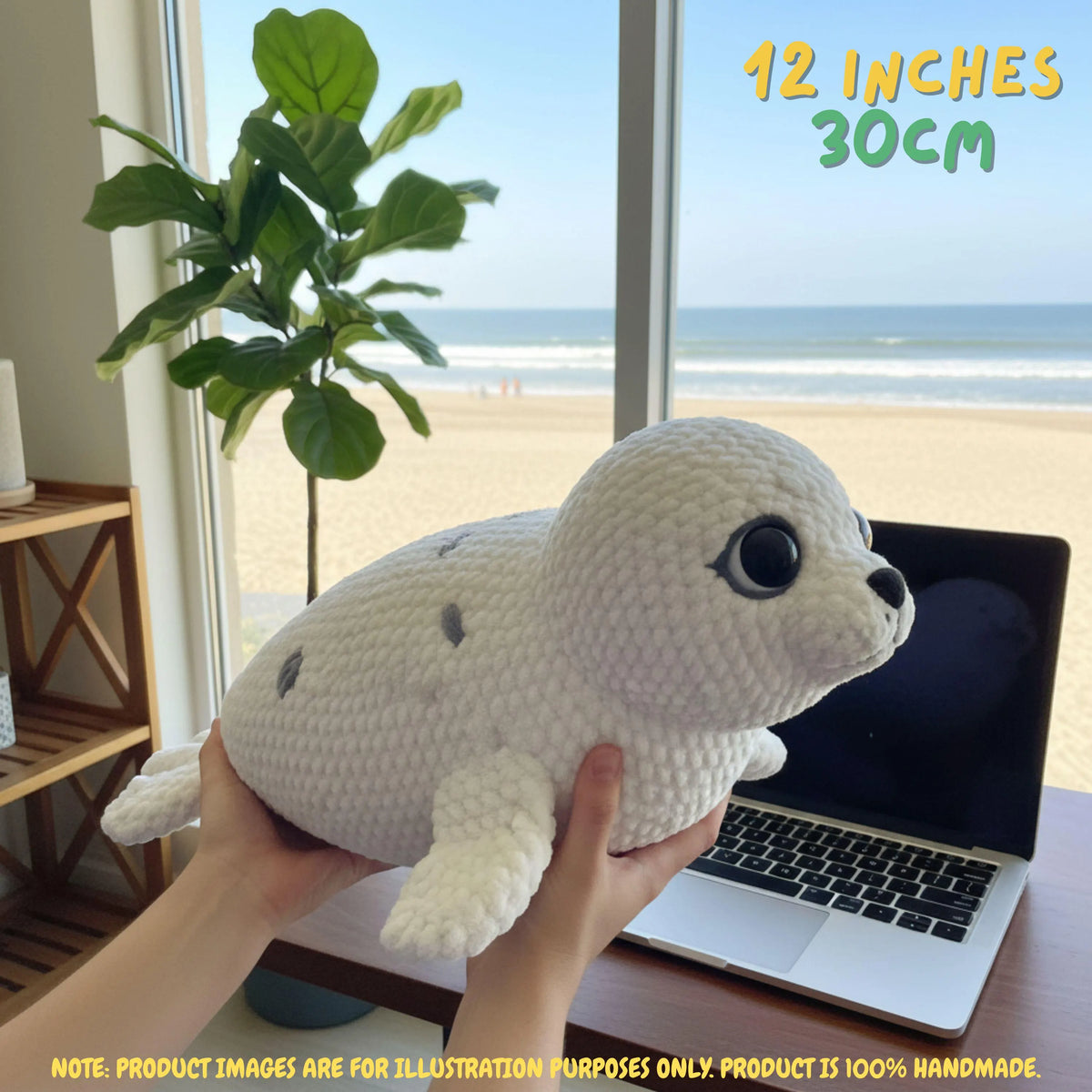 A crocheted seal plush toy being held by two hands in front of a window with a beach view. The plush is white with gray spots and large black eyes.