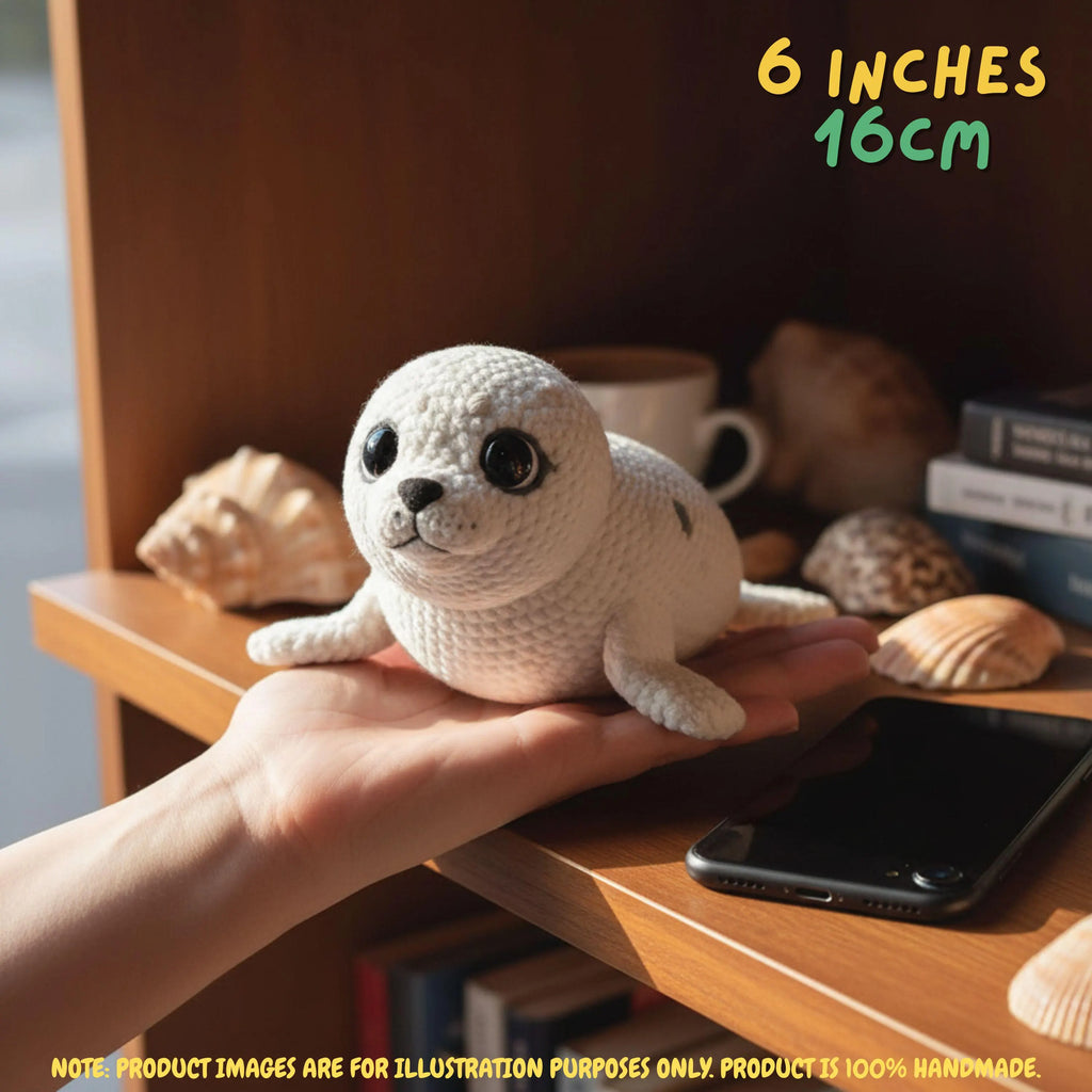 A small crocheted seal toy being held on a person's hand, placed on a wooden shelf with seashells, a mug, books, and a smartphone in the background. Text in the image reads '6 INCHES 16CM' and notes that the product is 100% handmade.