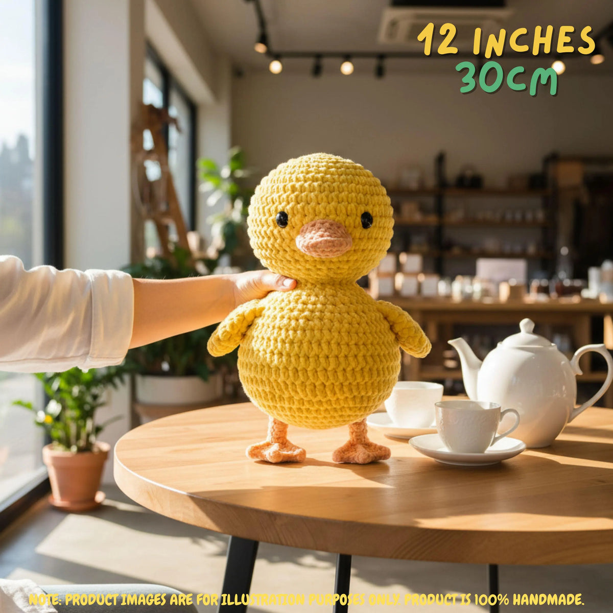 A yellow crocheted duck plush toy stands on a round wooden table in a cafe setting, with a person's arm reaching toward it. The text '12 INCHES 30CM' appears in the top right, and a note at the bottom says product images are for illustration purposes only and the product is 100% handmade.