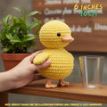 A yellow crocheted duck toy being held by a hand on a wooden surface, with a potted plant and a clear glass cup in the background. Text in the top right reads '6 INCHES 16CM', and a note at the bottom says the product image is for illustration purposes only and the product is 100% handmade.