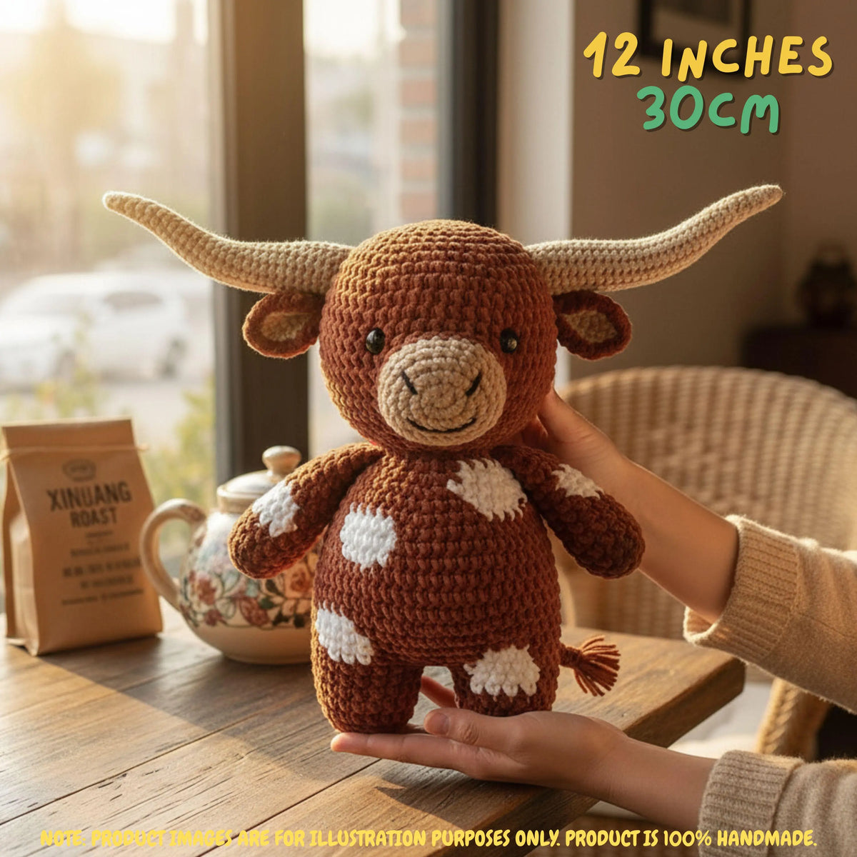 A crocheted longhorn bull plush toy being held by hands on a wooden table near a window, with a teapot and a bag labeled XINJIANG ROAST in the background. Text in the top right reads 12 INCHES 30CM.