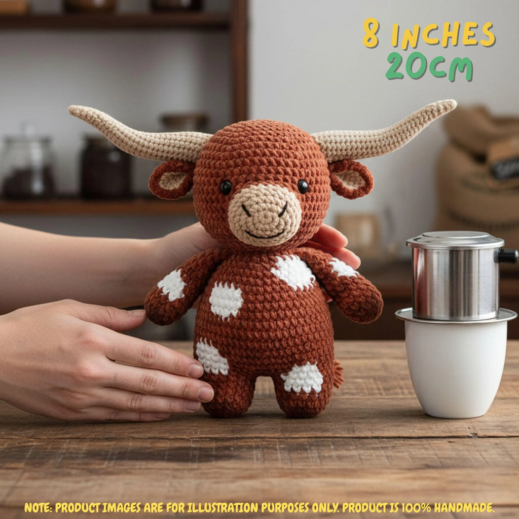 A crocheted plush toy bull with long cream horns and white spots on a brown body, held by hands on a wooden table with a coffee maker in the background. Text reads '8 INCHES 20CM' and includes a note that product images are for illustration purposes only.
