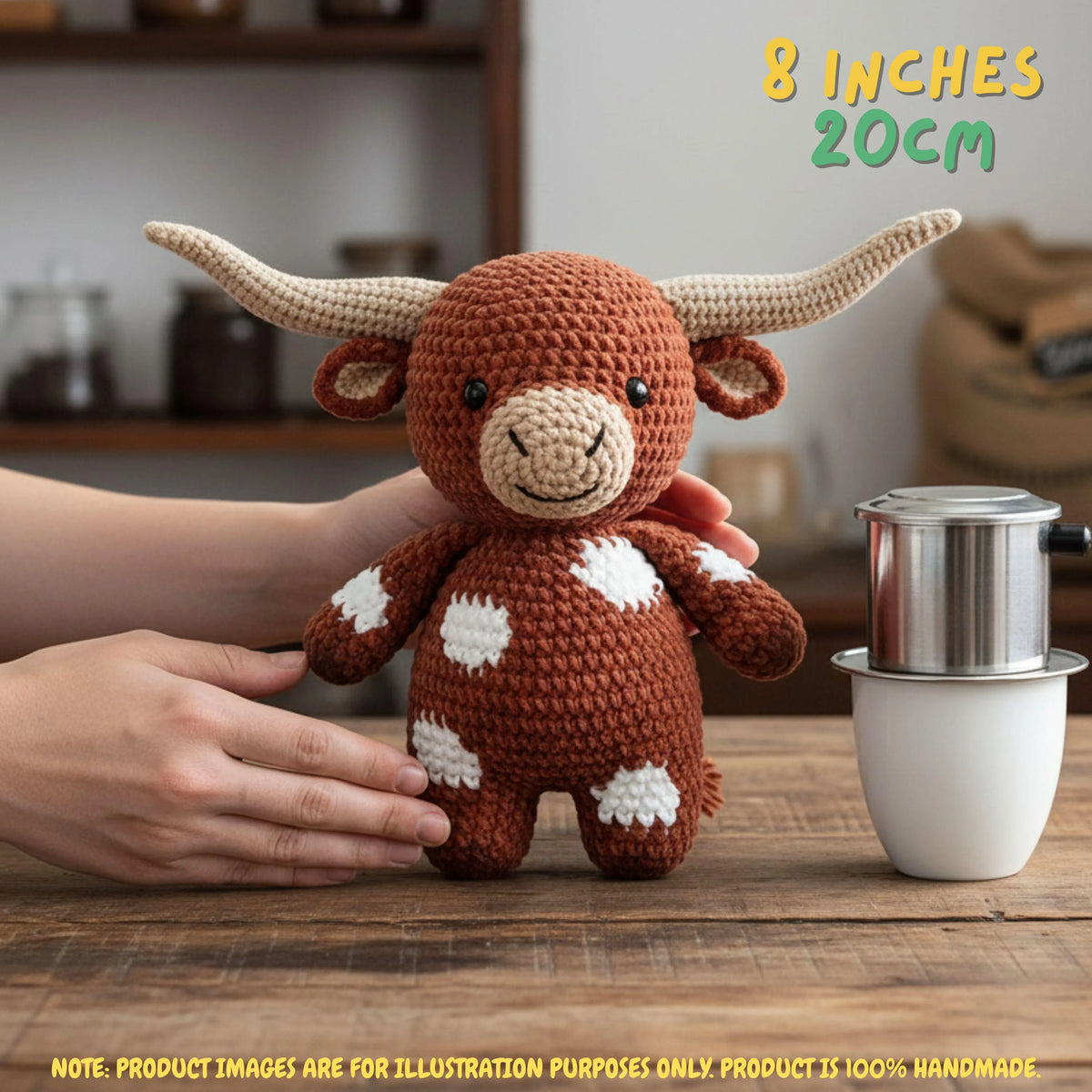 A crocheted plush toy bull with long cream horns and white spots on a brown body, held by hands on a wooden table with a coffee maker in the background. Text reads '8 INCHES 20CM' and includes a note that product images are for illustration purposes only.