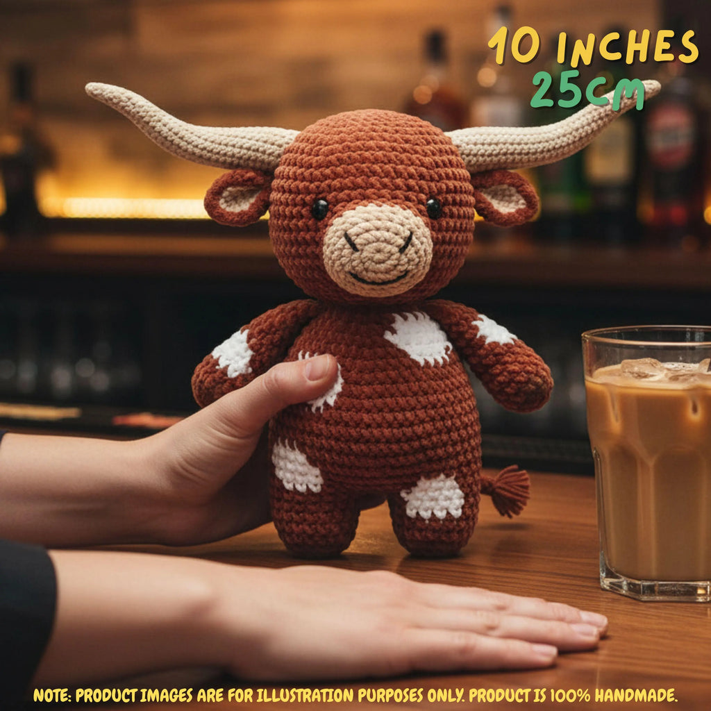 A crocheted toy bull with long cream horns, brown body, and white spots is held by two hands on a wooden table. A glass of iced coffee and a blurred bar background are visible behind it, with text overlays reading '10 INCHES 25CM' and a disclaimer at the bottom.