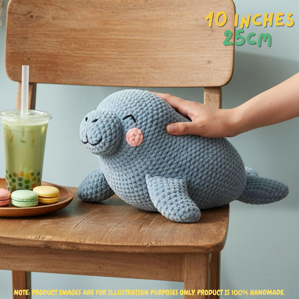 A crocheted seal plush toy in a dusty blue-gray color with a pink cheek and embroidered facial features, sitting on a wooden chair. A human hand is gently petting the plush.