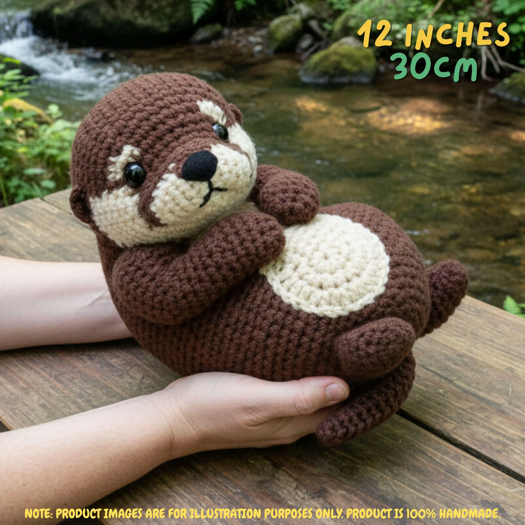 A crocheted otter plush toy being held in two hands on a wooden surface with a stream and greenery in the background. Yellow text in the top right reads '12 INCHES 30CM', and a note at the bottom says product images are for illustration purposes only and the product is 100% handmade.