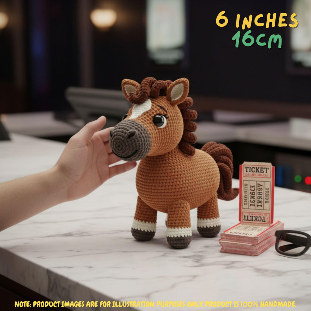 A crocheted toy horse in brown, cream, and dark brown is shown on a marble tabletop with a human hand touching its face. The background is a blurred indoor setting, and text overlays indicate the size as 6 inches (16 cm) and note that the product image is for illustration purposes only.