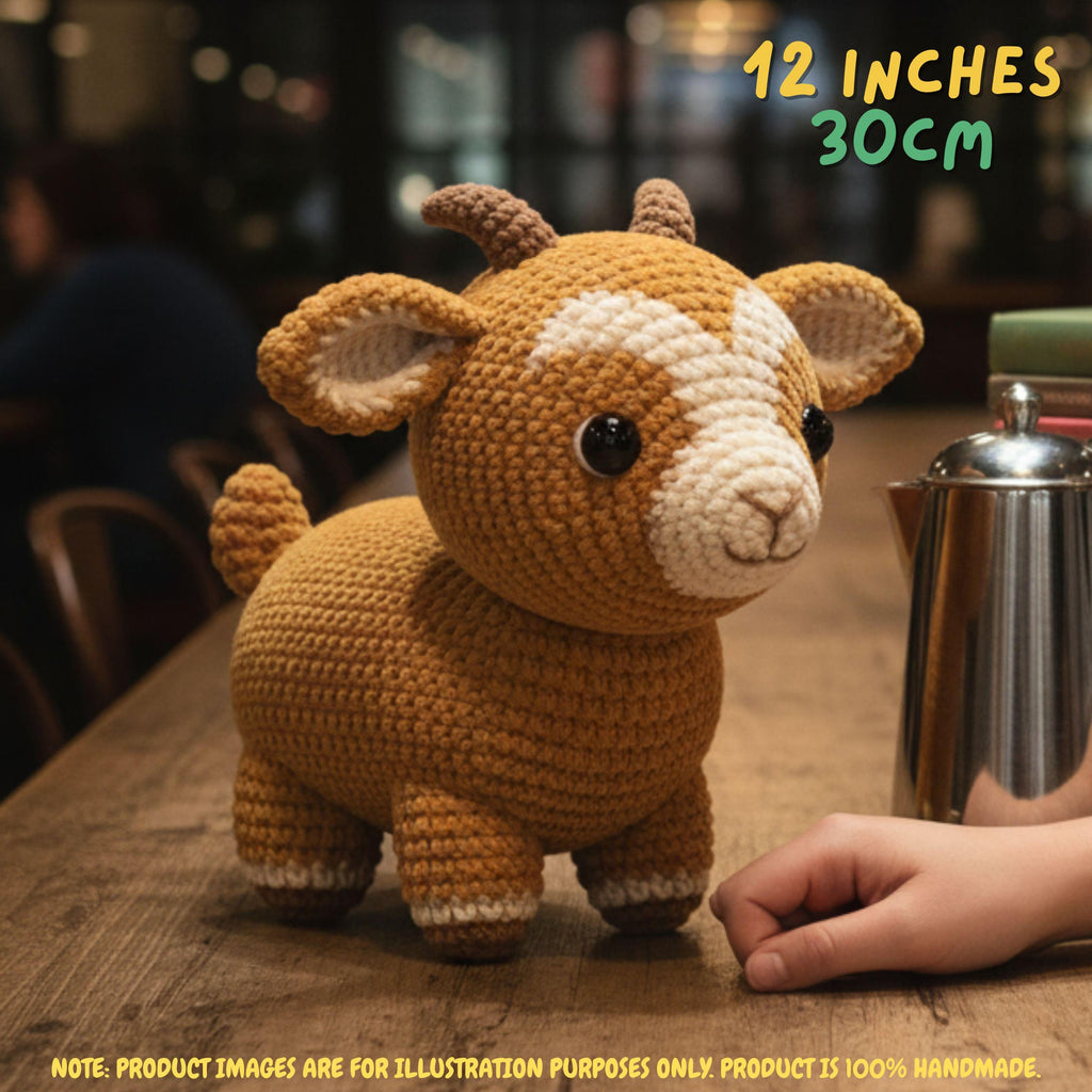 A crocheted stuffed goat toy sits on a wooden table in a warm indoor setting, with a human hand visible at the lower right for scale. Text in the top right reads '12 INCHES 30CM', and a note at the bottom says the product image is for illustration purposes only and the product is 100% handmade.