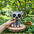 A crocheted raccoon toy being held by a human hand, placed on a small wooden slice outdoors with green foliage and a potted plant in the background. Text in the image reads '6 INCHES 16CM' and includes a note that product images are for illustration purposes only.