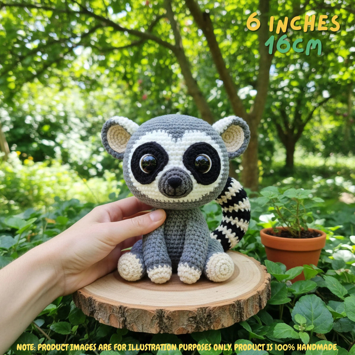A crocheted raccoon toy being held by a human hand, placed on a small wooden slice outdoors with green foliage and a potted plant in the background. Text in the image reads '6 INCHES 16CM' and includes a note that product images are for illustration purposes only.