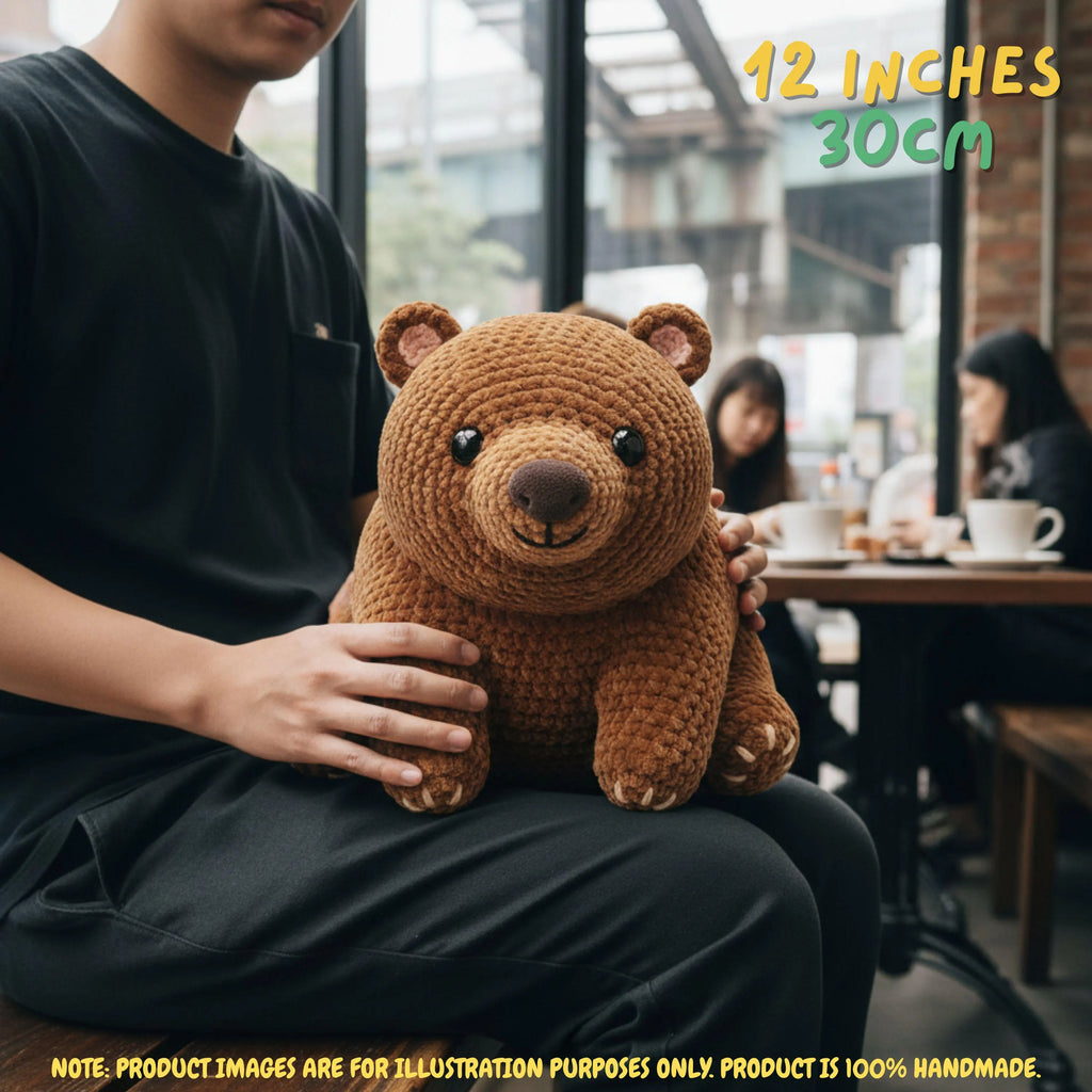 A person in a black shirt holds a brown crocheted teddy bear plush toy in a cafe setting. Text overlays indicate the size as 12 inches / 30cm, with a note at the bottom stating product images are for illustration purposes only.