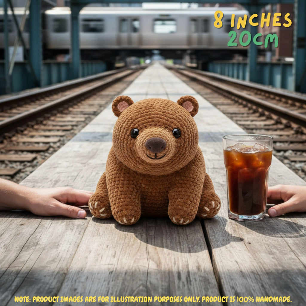 A crocheted brown teddy bear plush toy sits on a wooden train platform between two human hands, with a glass of iced coffee to its right and a blurred train and tracks in the background. Text overlays indicate the size as 8 inches / 20 cm.