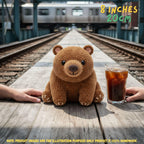 A crocheted brown teddy bear plush toy sits on a wooden train platform between two human hands, with a glass of iced coffee to its right and a blurred train and tracks in the background. Text overlays indicate the size as 8 inches / 20 cm.