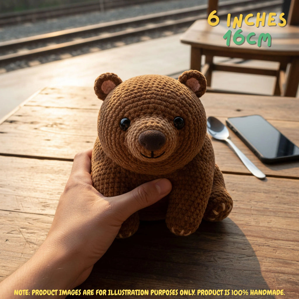 A small crocheted brown bear toy being held by a hand on a wooden table, with size text reading 6 inches / 16cm in the top right and a note at the bottom stating product images are for illustration purposes only.