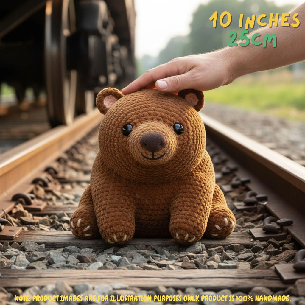 A brown crocheted teddy bear sits on railroad tracks, with a human hand resting on its head. The background shows blurred train tracks and greenery, with text overlays indicating the size is 10 inches (25 cm) and noting that the product is 100% handmade.