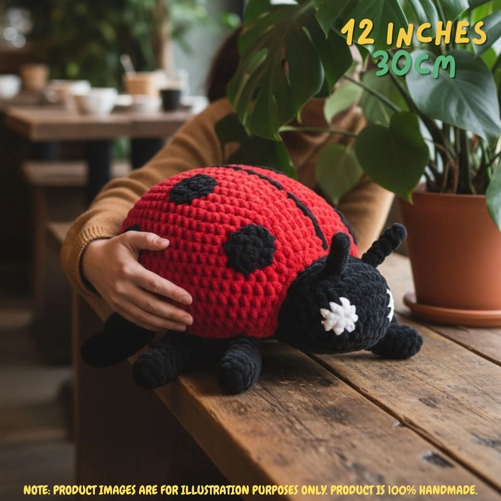 A crocheted ladybug plush toy being held by a person in a cozy indoor setting with a wooden table and potted plants. The text overlay indicates the size is 12 inches (30 cm) and notes that the product image is for illustration purposes only.