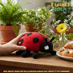 A crocheted ladybug toy being held by a hand on a wooden table, with potted plants, a small vase of flowers, and pastries in the background. Text in the top right reads '8 INCHES 20CM'.