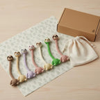 Collection of colorful long-neck handmade crochet sloth bag charms in brown, beige, purple, pink, and green lined up on a patterned cloth with a gift box and drawstring pouch.