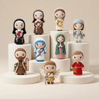A collection of crocheted religious doll figurines displayed on white cylindrical pedestals against a warm beige background. The dolls represent various saints and angels, each with distinct colorful garments and accessories.