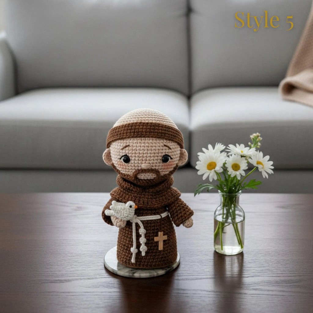 A crocheted doll of a Franciscan monk holding a white dove stands on a small round base on a dark wooden table, next to a small glass vase with white daisies. The background shows a blurred gray sofa with a beige throw blanket.