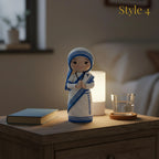 A crocheted female figurine styled on a wooden bedside table next to a lit white lamp, a glass cup, and a book, in a softly blurred bedroom setting. The figurine is white with blue stripes and a black face, and the text 'Style 4' appears in the top right corner.