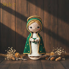 A crocheted doll styled as a robed figure with a green hooded cloak, cream dress, and gold trim, standing on a wooden surface with dried flowers and a dark wood plank background.