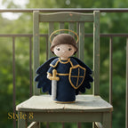 Crocheted angel doll wearing a dark blue robe with gold trim, a brown helmet, and holding a sword and shield, displayed on a rustic green wooden chair with a blurred garden background.