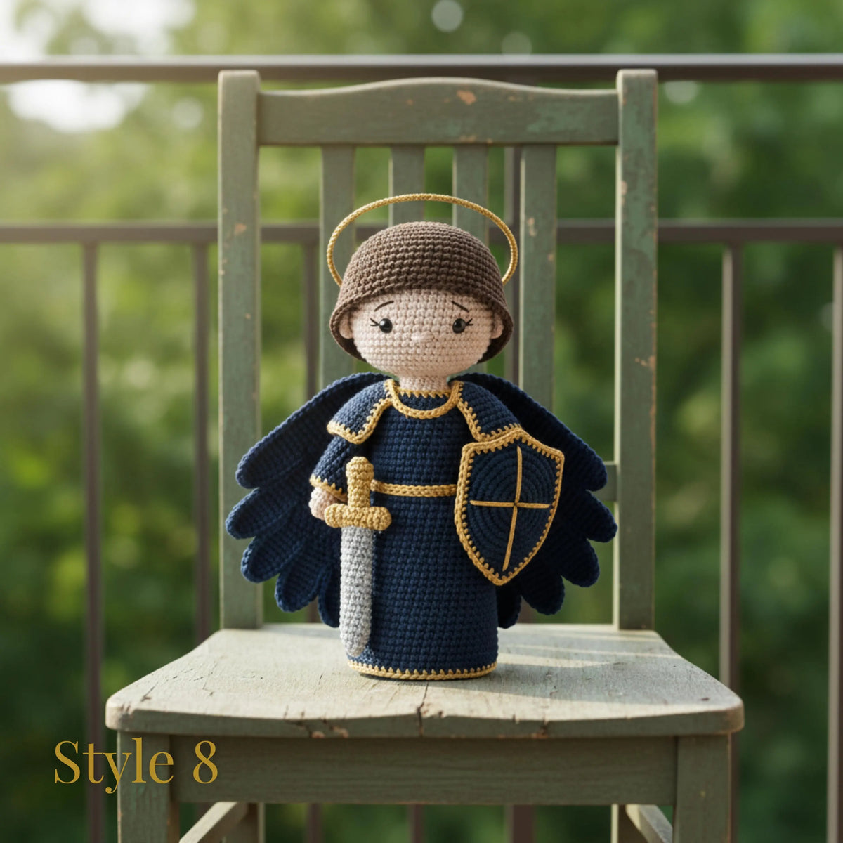 Crocheted angel doll wearing a dark blue robe with gold trim, a brown helmet, and holding a sword and shield, displayed on a rustic green wooden chair with a blurred garden background.